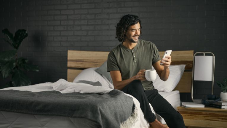 person is sitting on a bed smiling at their phone while holding mug