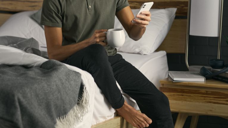 Person sitting on a bed holding a white mug and smartphone